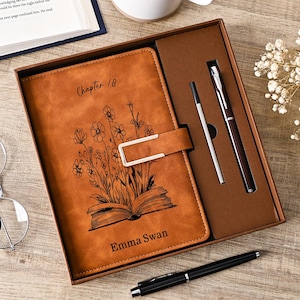 May include: A tan leather-like journal set in a brown box, featuring a metal clasp, a black line drawing of flowers emerging from an open book, and the text "Chapter 18" and "Emma Swan". The set includes a matching pen and a silver ink refill.