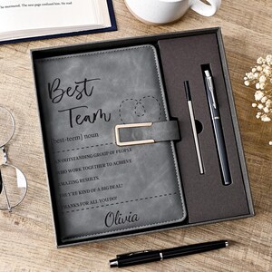 Puede incluir: Un cuaderno gris con un cierre metálico dorado, con el texto Best Team, una definición y el nombre Olivia. El cuaderno se presenta en una caja de regalo gris oscuro con un bolígrafo negro y un recambio, más un bolígrafo negro debajo.