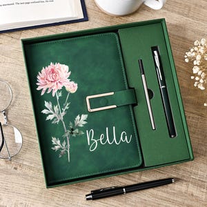 May include: A dark green notebook set in a matching box, featuring a pink chrysanthemum illustration and the name Bella in white script. The set includes a black pen and a refill cartridge, with a gold-toned metal clasp on the notebook cover.