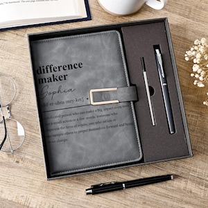 Könnte beinhalten: Ein graues Notizbuch mit den Worten "difference maker Sophia" und einer Definition, präsentiert in einer Geschenkbox mit einem Stift. Das Notizbuch hat einen goldfarbenen Verschluss. Ein zweiter Stift liegt auf der Holzoberfläche.