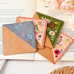 May include: Four triangular corner bookmarks made of felt and paper. Each bookmark features hand-stitched embroidery with floral motifs and a single letter: N on grey, A on light peach, Q on dark green, and L on light peach felt.