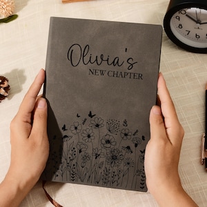Op de afbeelding: Een grijs notitieboek met de tekst "Olivia's NEW CHAPTER" en een bloemenontwerp. Een zwarte wekker, een pen en decoratieve items zijn ook zichtbaar.
