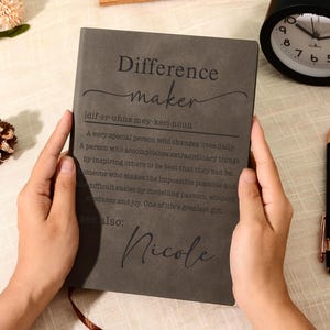 Puede incluir: Un cuaderno gris oscuro con el texto Difference maker, una guía de pronunciación fonética y una definición impresa en la portada. El nombre Nicole está impreso en la parte inferior. El cuaderno es sostenido por dos manos, con un reloj y bolígrafo.