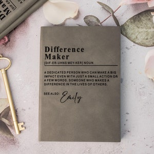 Puede incluir: Un cuaderno gris con el título "Difference Maker" impreso en la portada. La definición de "Difference Maker" está impresa debajo. Una llave dorada y un libro también están en la imagen.