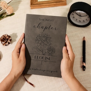 May include: A gray journal with the words "THE NEXT chapter" and "ELOUlSE" engraved on the cover, along with a floral design. A black alarm clock and pen are also visible.