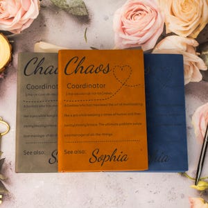 May include: Three notebooks with the words "Chaos Coordinator" printed on them. The notebooks are in shades of grey, orange, and blue. The orange notebook has the name "Sophia" printed on it.