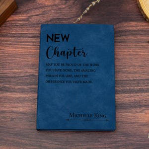 Puede incluir: Un diario de imitación de cuero azul con el texto "NEW Chapter" y la cita "May you be proud of the work you have done, the amazing person you are, and the difference you have made." El diario está personalizado con el nombre "Michelle King".