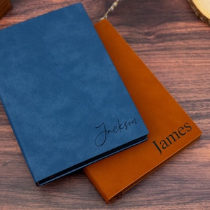 May include: Two personalised notebooks, one blue and one orange, are shown on a wooden surface. The blue notebook has the name "Jackson" in cursive, while the orange notebook has "James" in a serif font.