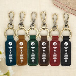 May include: Six rectangular leather keychains in teal, tan, green, brown, red, and black. Each features a white Spotify logo and a sound wave graphic printed on the front, attached to a bronze-toned metal swivel lobster clasp and a split ring.