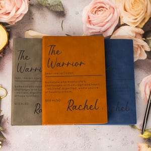 May include: Three hardback journals in grey, orange, and blue. Each journal features the text "The Warrior" and a definition. The orange journal is personalised with the name "Rachel". The journals are surrounded by pink and peach roses.