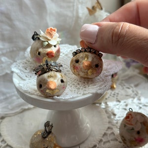 Puede incluir: Una colección de adornos de pájaros caprichosos y hechos a mano. Cada pájaro presenta un diseño único, con detalles de pan de oro, rasgos faciales delicados y adornado con coronas en miniatura o adornos florales. Los pájaros se exhiben sobre un pedestal blanco y tapetes de encaje.