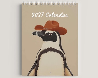 May include: A spiral-bound 2027 calendar featuring a graphic illustration of a penguin wearing a brown cowboy hat. The penguin is depicted against a solid light tan background with the white text 2027 Calendar printed at the top center.