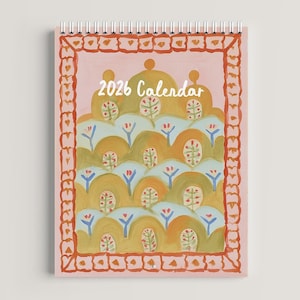 May include: A spiral-bound 2026 calendar with a whimsical, painted design. The cover features a pink background with a decorative border and a landscape of stylized hills in shades of yellow, green, and orange. The text "2026 Calendar" is in white.