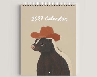 May include: A spiral-bound 2027 calendar with a light tan cover featuring an illustration of a black and white skunk wearing a brown cowboy hat. The text 2027 Calendar is printed in white at the top center of the cover above the skunk illustration.
