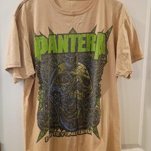 May include: A tan short-sleeve t-shirt featuring a graphic print of a skull with green and blue details. The word PANTERA is printed in large green letters above the skull, and the text FAR BEYOND DRIVEN is printed in smaller letters below the skull.
