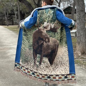 May include: A long denim jacket with a moose tapestry on the back. The jacket has a sherpa lining and a fringe trim.