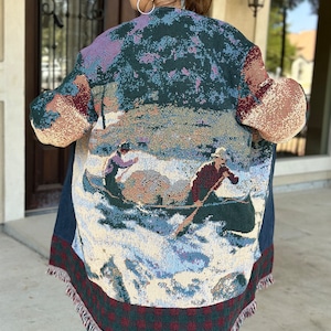 May include: A long, green, blue, and red tapestry cardigan with a fringed hem. The cardigan features a woven design of a winter scene with people ice skating.