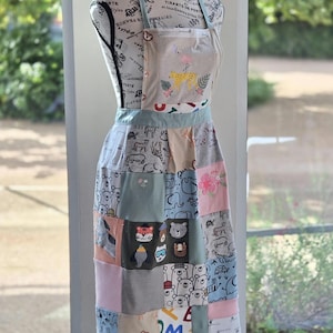 May include: A patchwork apron with a light blue neck strap and waist tie. The top section features a flamingo and leopard print. The lower section is a patchwork of animal and floral prints in various colors, including pink, blue, and gray. The apron is displayed on a mannequin.