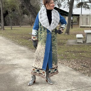 May include: A long blue denim coat with a faux fur collar and a colorful patterned design. The coat has fringe trim at the hem and is worn with snake print boots.