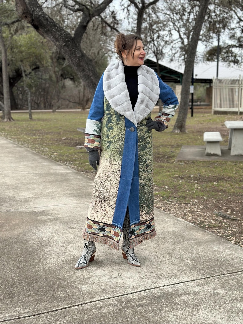 May include: A long blue denim coat with a faux fur collar and a colorful patterned design. The coat has fringe trim at the hem and is worn with snake print boots.