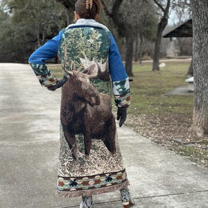 May include: A long denim jacket with a moose tapestry design on the back. The jacket has a fringed hem and a brown faux fur collar.