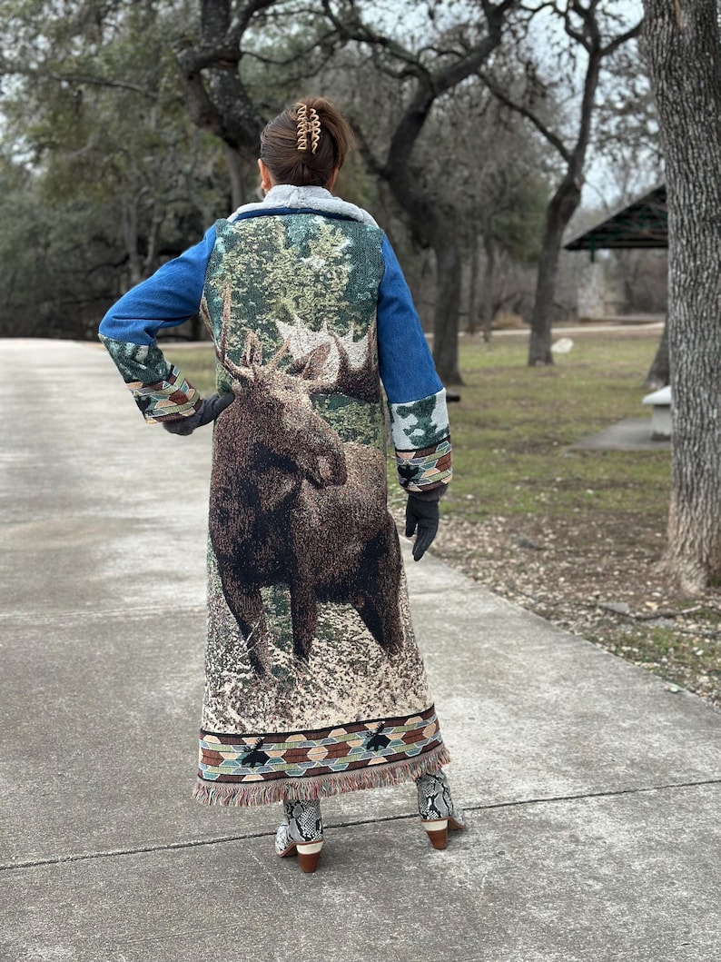 May include: A long denim jacket with a moose tapestry design on the back. The jacket has a fringed hem and a brown faux fur collar.