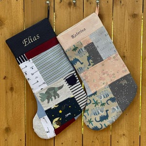 May include: Two personalized Christmas stockings hanging on a wooden fence. One stocking is navy blue with the name "Elias" embroidered in white. The other is pink with the name "Katerina". Both stockings are patchwork with various fabric patterns.