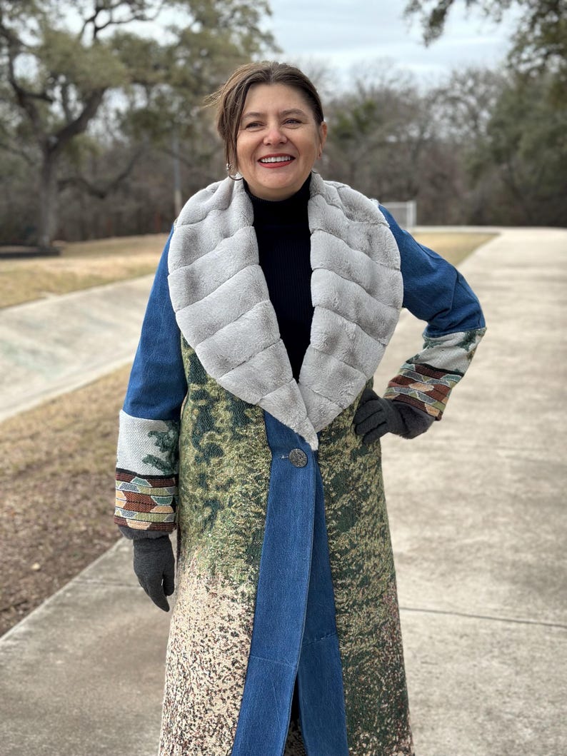 May include: A long blue denim coat with a faux fur collar and a green and brown patterned design. The coat has a button closure and a long sleeve.