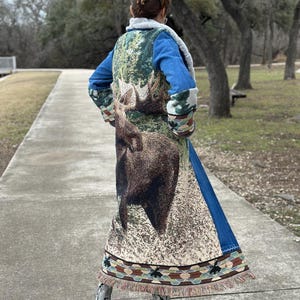 May include: A long denim jacket with a tapestry back featuring a moose design. The tapestry is in shades of green, brown, and beige. The jacket has a fringe trim.