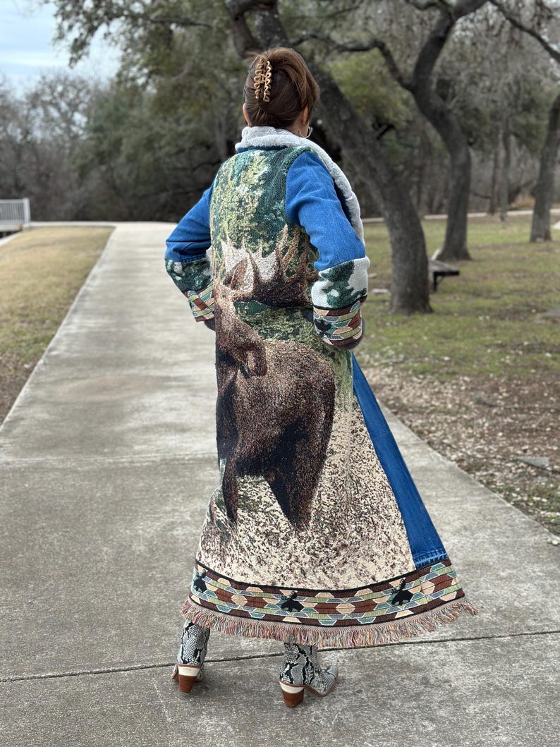 May include: A long denim jacket with a tapestry back featuring a moose design. The tapestry is in shades of green, brown, and beige. The jacket has a fringe trim.
