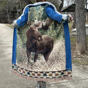 May include: A long denim jacket with a tapestry back featuring a moose in a forest scene. The tapestry has a colorful fringe border.
