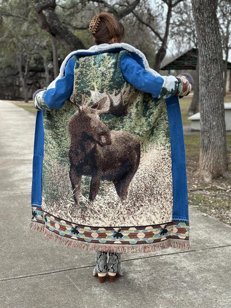 May include: A long denim jacket with a tapestry back featuring a moose in a forest scene. The tapestry has a colorful fringe border.
