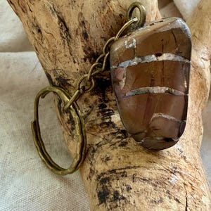 May include: A keychain featuring a polished, irregular brown stone with white veined patterns, attached to a brass-toned metal chain and a circular split ring. The stone has a smooth, glossy finish and is connected to the hardware by a small loop.
