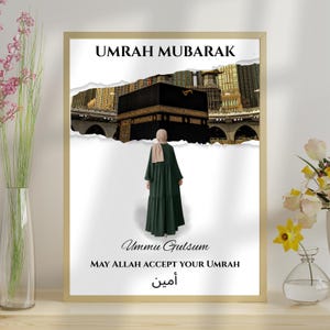 May include: A framed print with a white background and a torn paper effect. The image shows a woman in a green abaya standing in front of the Kaaba in Mecca. The text on the print reads "Umrah Mubarak", "Ummu Gulsom", "May Allah accept your Umrah", and "آمين".