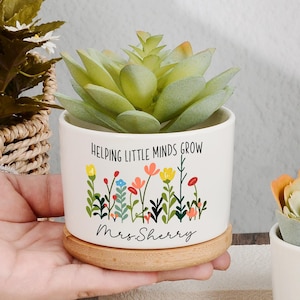 May include: Two white ceramic cylindrical planters with light wood saucers, each containing a faux succulent. One planter features a colourful wildflower print with the text "Helping Little Minds Grow" and "Mrs. Sherry". The other displays a floral design with "Mrs. Elaine".