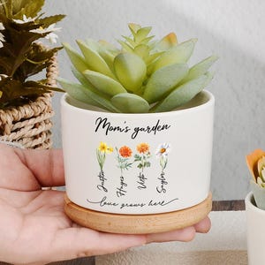 May include: Two white ceramic planters with faux succulents, each on a wooden base. The planters are inscribed with "Mom's garden" and "Love grows here," along with names. One planter is held by a hand. The plants have green and orange hues.
