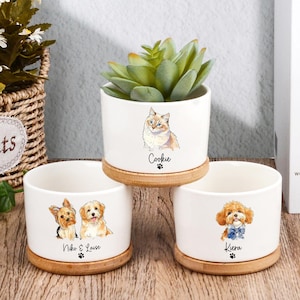 Könnte beinhalten: Weiße Keramik-Pflanztöpfe mit Aquarellporträts von Haustieren. Die Töpfe zeigen eine Katze und zwei Hunde, jeweils mit Namen und Pfotenabdruck. Die Töpfe stehen auf Bambusuntersetzern, mit einer Sukkulente in einem.