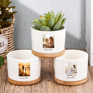 May include: Three white ceramic planters with bamboo bases, each featuring a photo and the text "Love you Mom." One planter holds a succulent. The photos depict a mother and child. The planters are on a wooden surface.