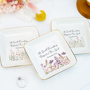May include: Three square white ceramic trinket dishes with gold-toned rims. Each dish features a floral illustration and the black printed text A Sweet Friendship Refreshes The Soul Proverbs 27:9. A silver ring sits near the dishes.