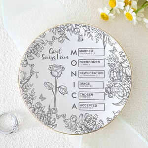 Può includere: Un piattino rotondo in ceramica bianca con bordo dorato, decorato con disegni floreali neri, una rosa singola e il testo God Says I Am MONICA con sei riquadri contenenti versetti biblici: Marked, Overcomer, New Creation, Image, Chosen e Accepted.