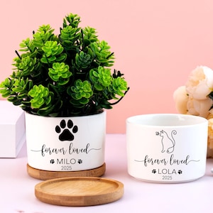 May include: Two white ceramic pet bowls with the words "forever loved" and a pet's name and year. One bowl has a paw print, the other a cat silhouette. A small green plant in a white pot with a paw print design sits on a wooden coaster.
