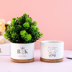 May include: Two white ceramic planters with bamboo bases. The planter on the left has a black and white design with the initials "R & K" and the date "08.10.2024". The planter on the right has a black and white design of two hands holding each other with the text "James & Amelia 01.29.2024".