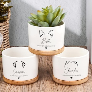 Peut inclure: Trois pots de fleurs cylindriques en céramique blanche avec soucoupes en bambou. Chaque pot présente une illustration noire d'oreilles de chien, un nom et le texte "FOREVER IN OUR HEARTS" avec un petit cœur. Un pot contient une succulente.