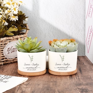 Puede incluir: Dos macetas de cerámica blanca con suculentas, cada una sobre una base de madera. Las macetas están inscritas con "James + Sophia married 09.02.2025" y un diseño botánico. Una maceta tiene suculentas verdes, la otra tiene suculentas naranjas y verdes.