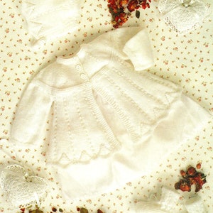 Könnte beinhalten: Ein weißer gestrickter Baby-Cardigan mit Knopfleiste und einer passenden weißen Strickmütze. Der Cardigan hat einen gewellten Saum und liegt auf einem floral gemusterten Stoff.