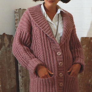 May include: A long-sleeved, pink cardigan with a chunky knit pattern, featuring a large collar, button closure, and two pockets. The cardigan is made with a soft, textured yarn.
