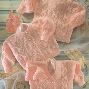 Może przedstawiać: Kolekcja trzech jasnoróżowych swetrów i kardiganów. Swetry mają wzór o fakturze i są ozdobione małymi guzikami. Ubrania są prezentowane na jasnoniebieskiej drewnianej powierzchni, z dekoracyjną lalką i nutami w tle.