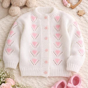 May include: A white knitted baby cardigan featuring pink heart patterns and pink buttons. The image also shows pink baby shoes with bows. Other baby accessories, including a plush bunny, are also present.