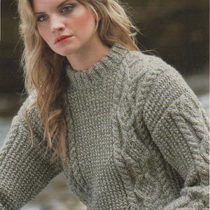 May include: A close-up of a woman wearing a light green cable knit jumper. The jumper has a round neckline and long sleeves. The woman has long blonde hair and is wearing jeans.