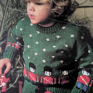May include: A green knitted jumper with a white snowflake pattern and a red and blue pattern of toy soldiers. The jumper has long sleeves and a crew neck.